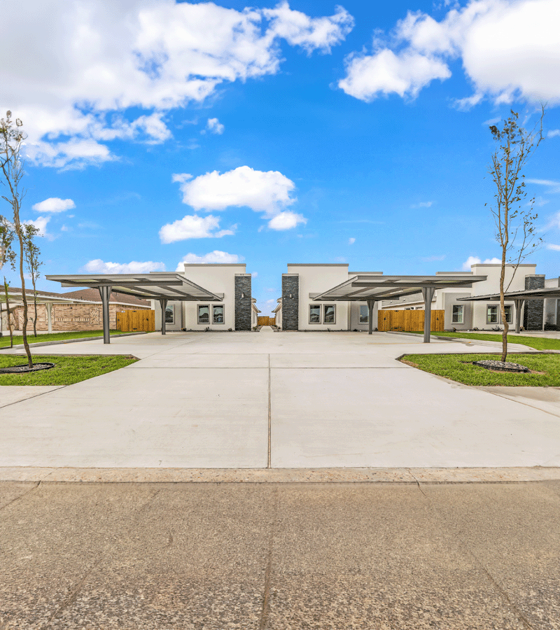 Two duplex units facing a shared concrete drive, each with a carport and newly planted trees